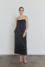 Front view of a model wearing the black silk charmeuse Kamperett Agnes Slip Dress, a boxy midi slip with straight neckline and corded straps.