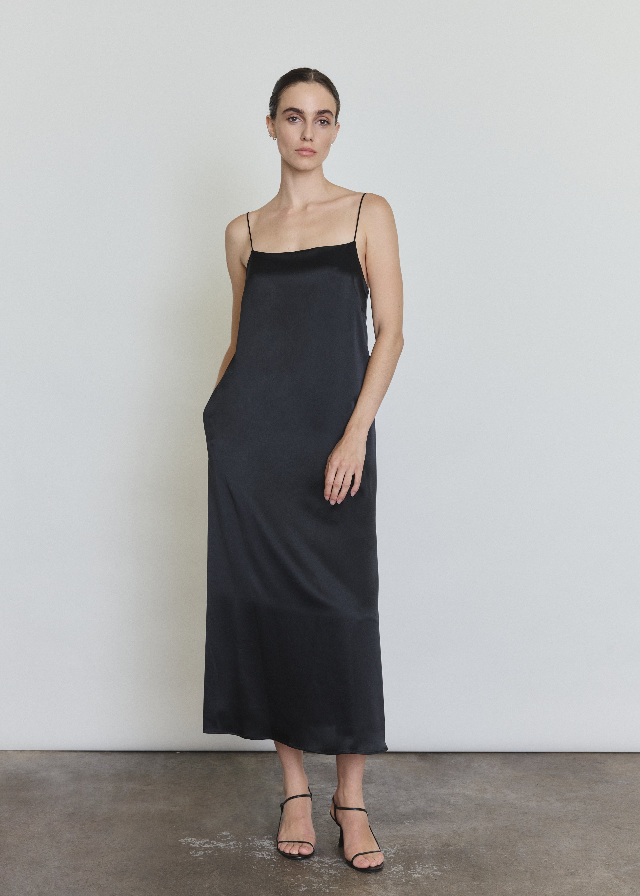 Front view of a model wearing the black silk charmeuse Kamperett Agnes Slip Dress, a boxy midi slip with straight neckline and corded straps.