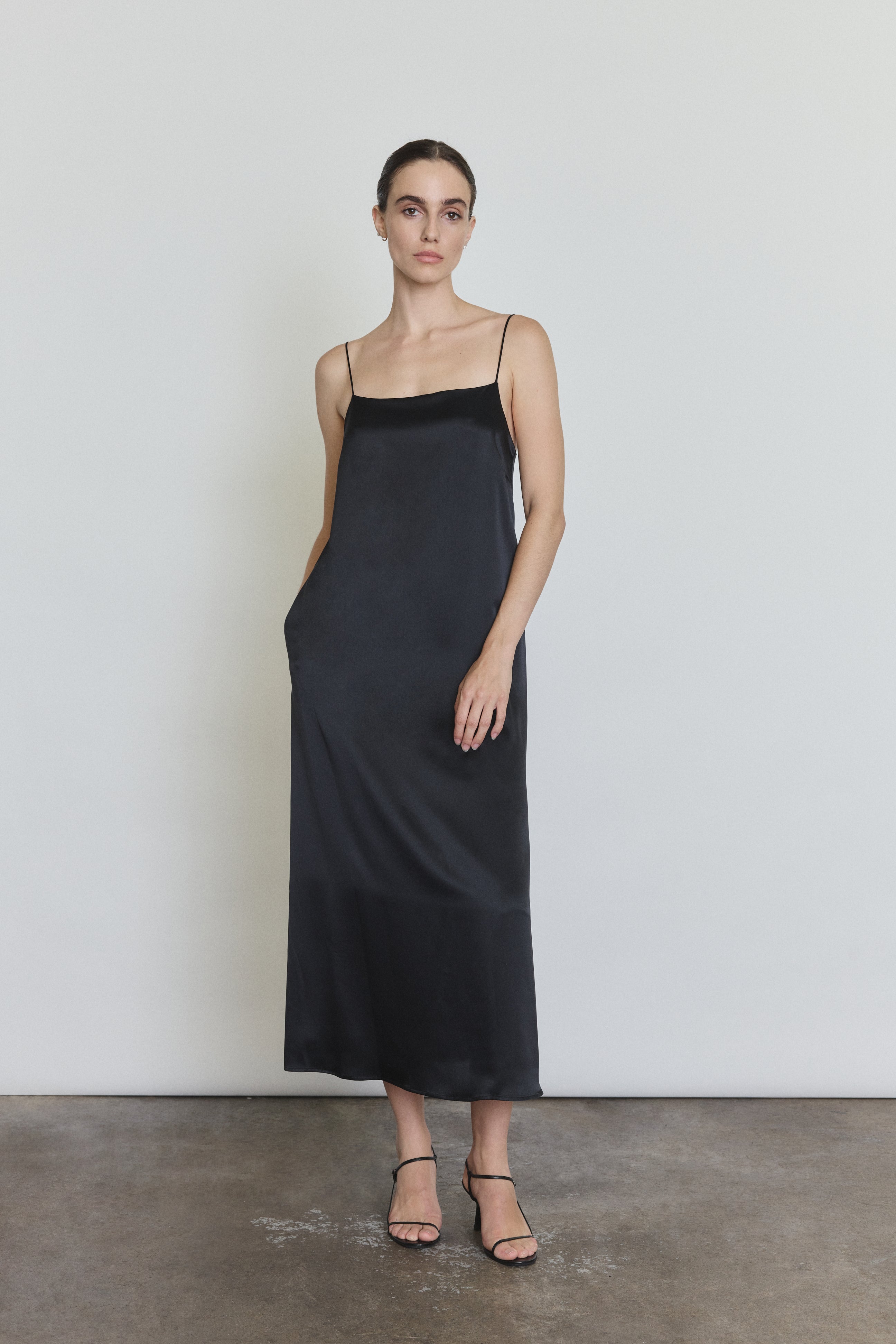 Front view of a model wearing the black silk charmeuse Kamperett Agnes Slip Dress, a boxy midi slip with straight neckline and corded straps.
