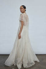 Left profile of ivory silk organza Anemoia Gown. Clean column silhouette with metallic lace detailing and deconstructed raw hem cap sleeve.