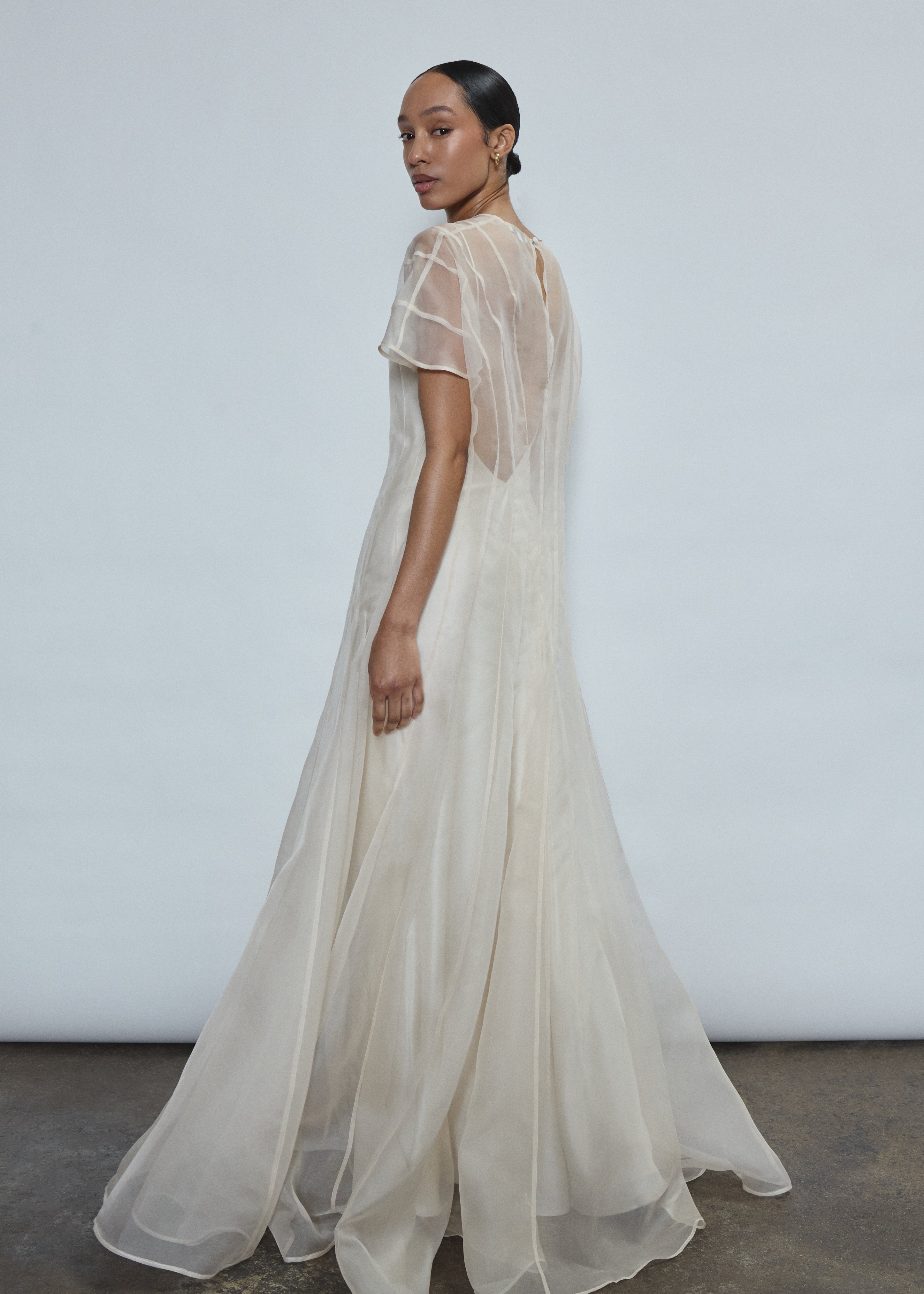 Left profile of ivory silk organza Anemoia Gown. Clean column silhouette with metallic lace detailing and deconstructed raw hem cap sleeve.