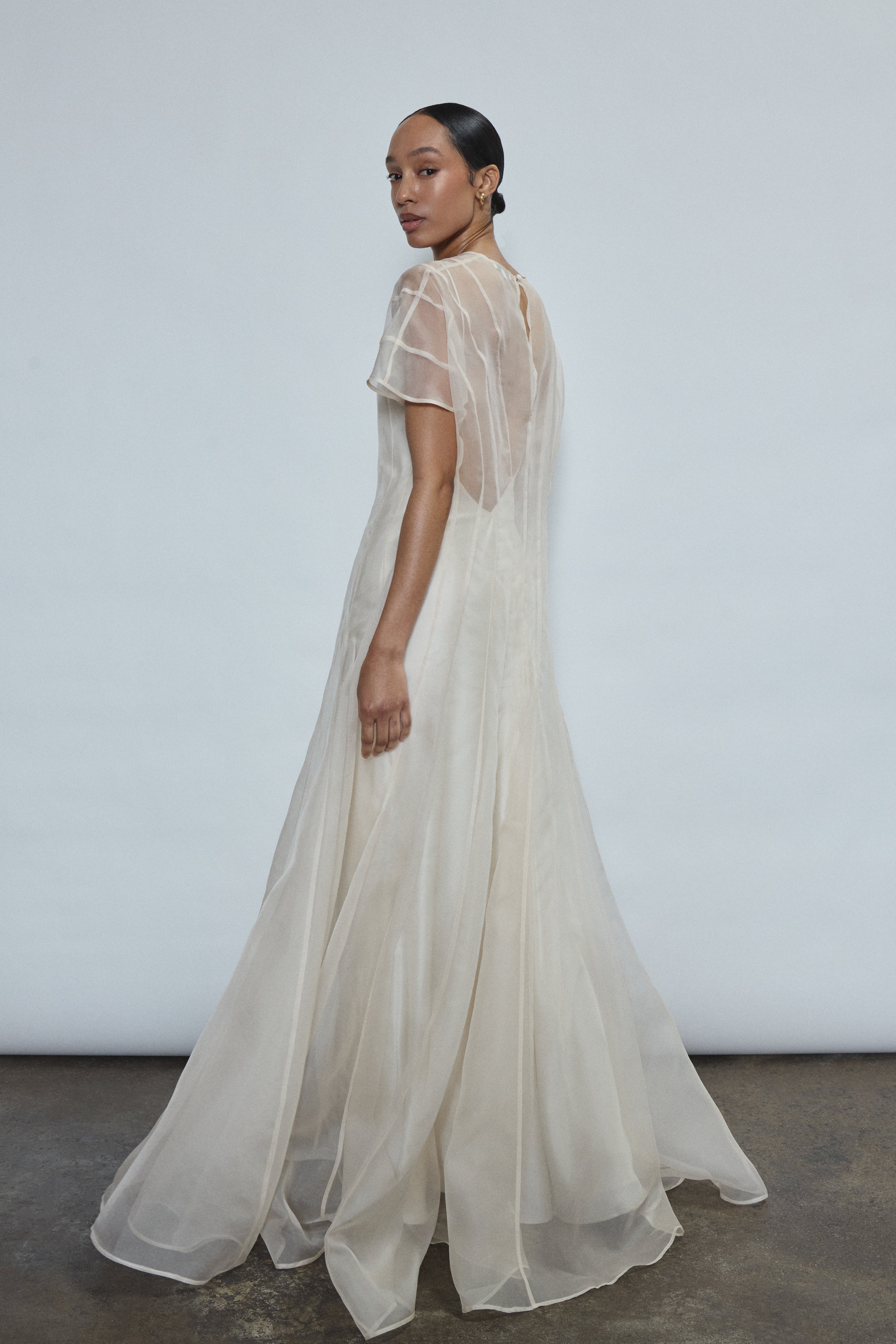 Left profile of ivory silk organza Anemoia Gown. Clean column silhouette with metallic lace detailing and deconstructed raw hem cap sleeve.