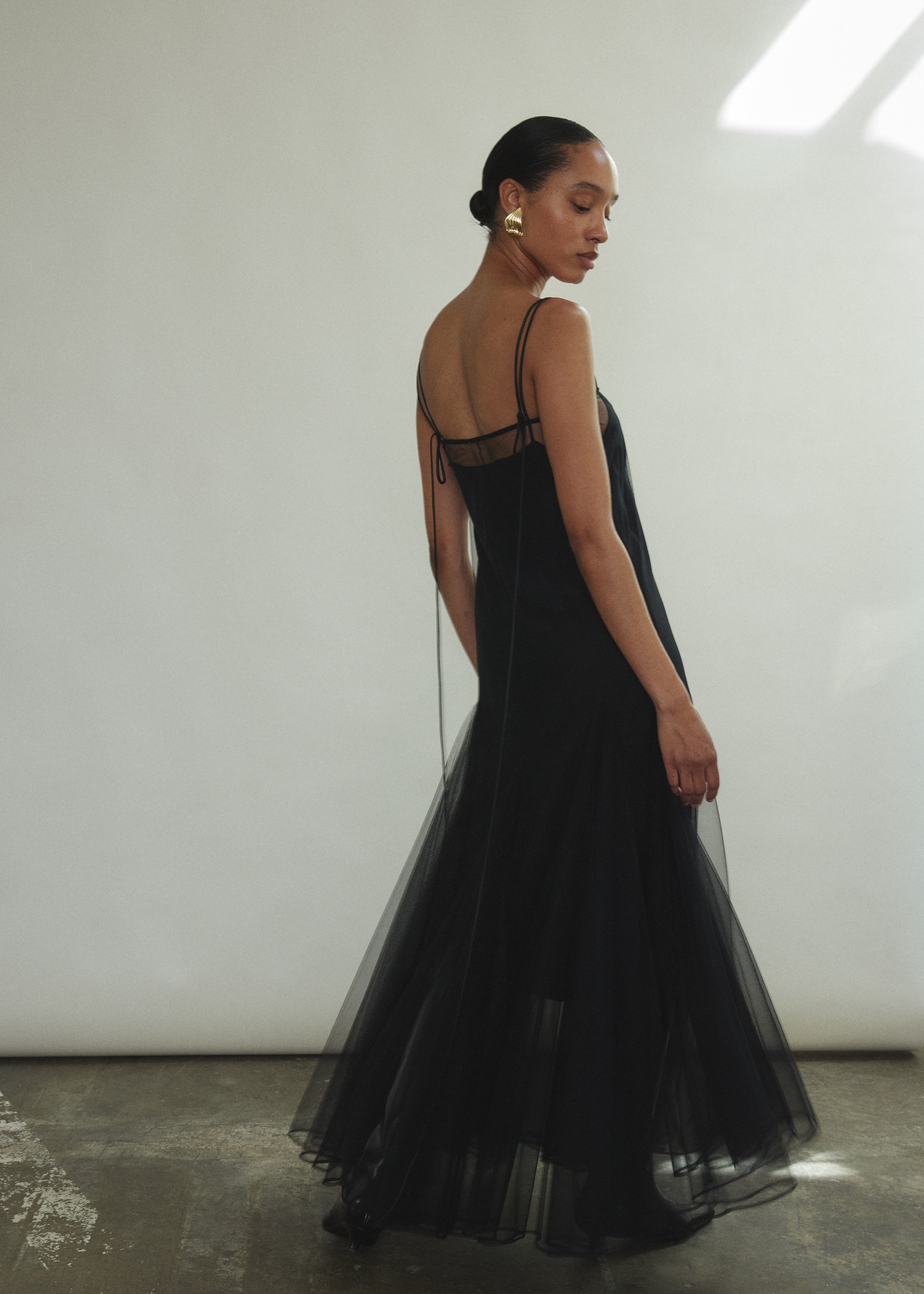 Model wearing the brown Kamperett Camille Dress, a sheer tulle tea-length overlay featuring adjustable silk corded straps and a voluminous godet-pleated skirt.