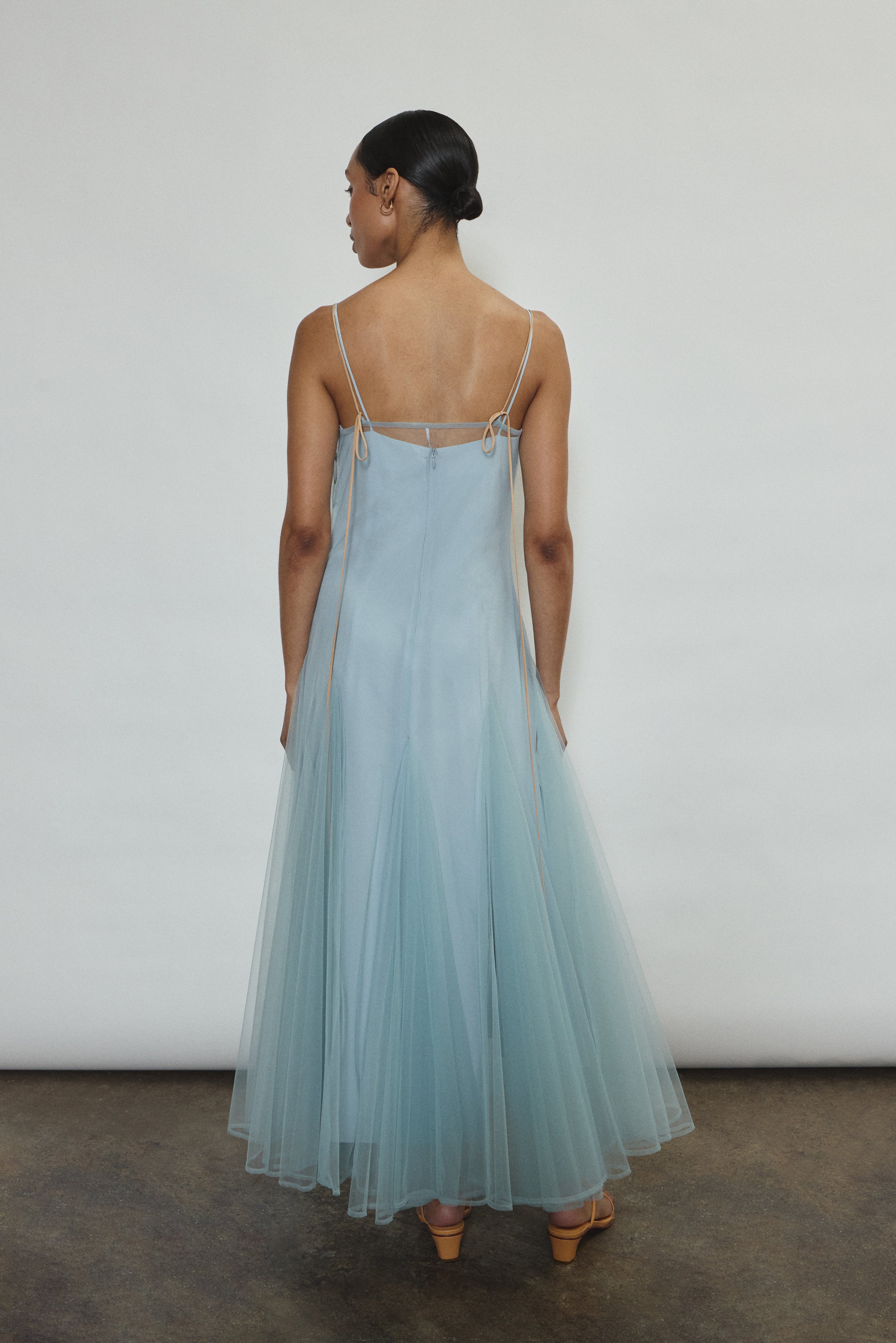 Model wearing the blue Kamperett Camille Dress, a sheer tulle tea-length overlay featuring adjustable silk corded straps and a voluminous godet-pleated skirt.