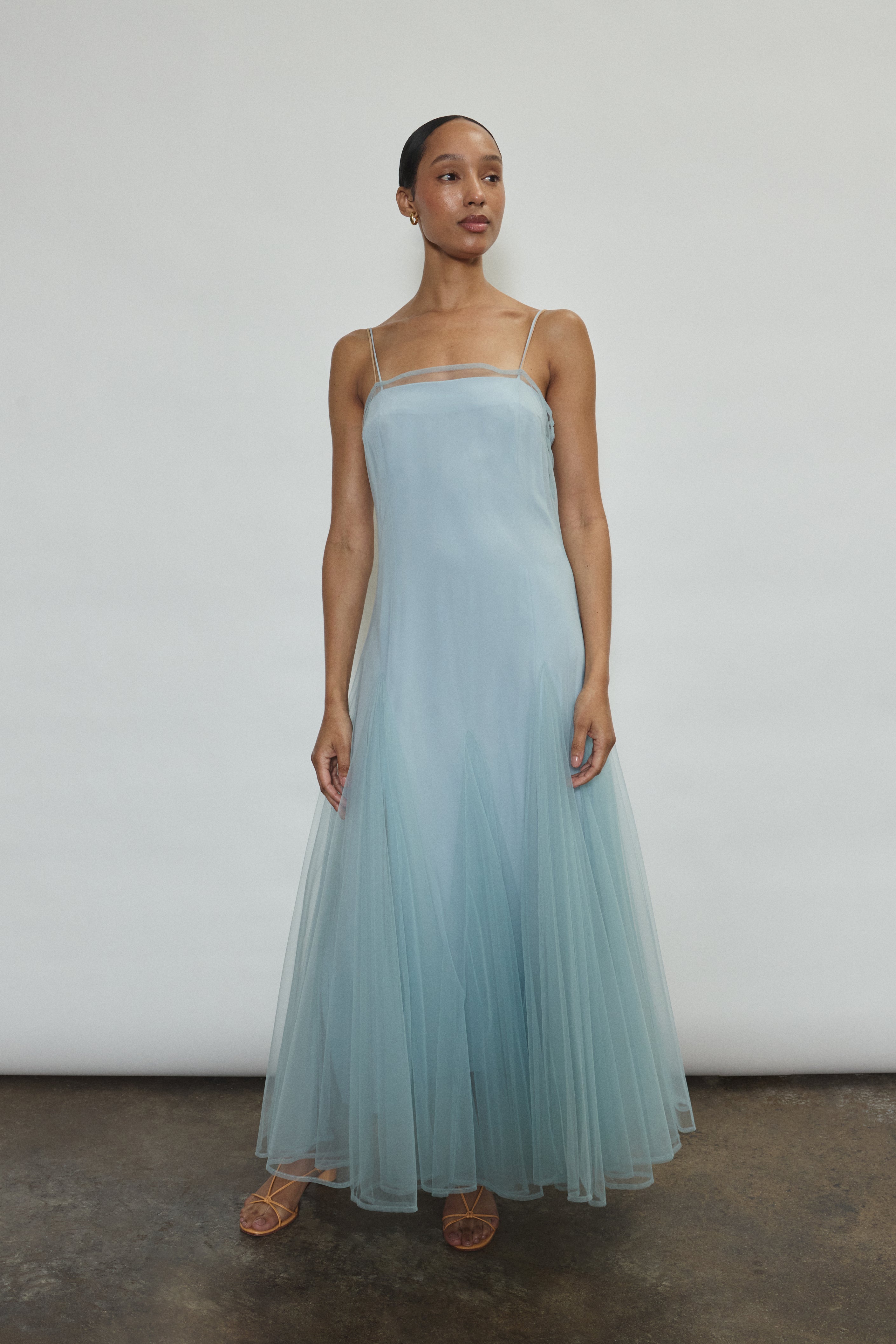 Model wearing the blue Kamperett Camille Dress, a sheer tulle tea-length overlay featuring adjustable silk corded straps and a voluminous godet-pleated skirt.