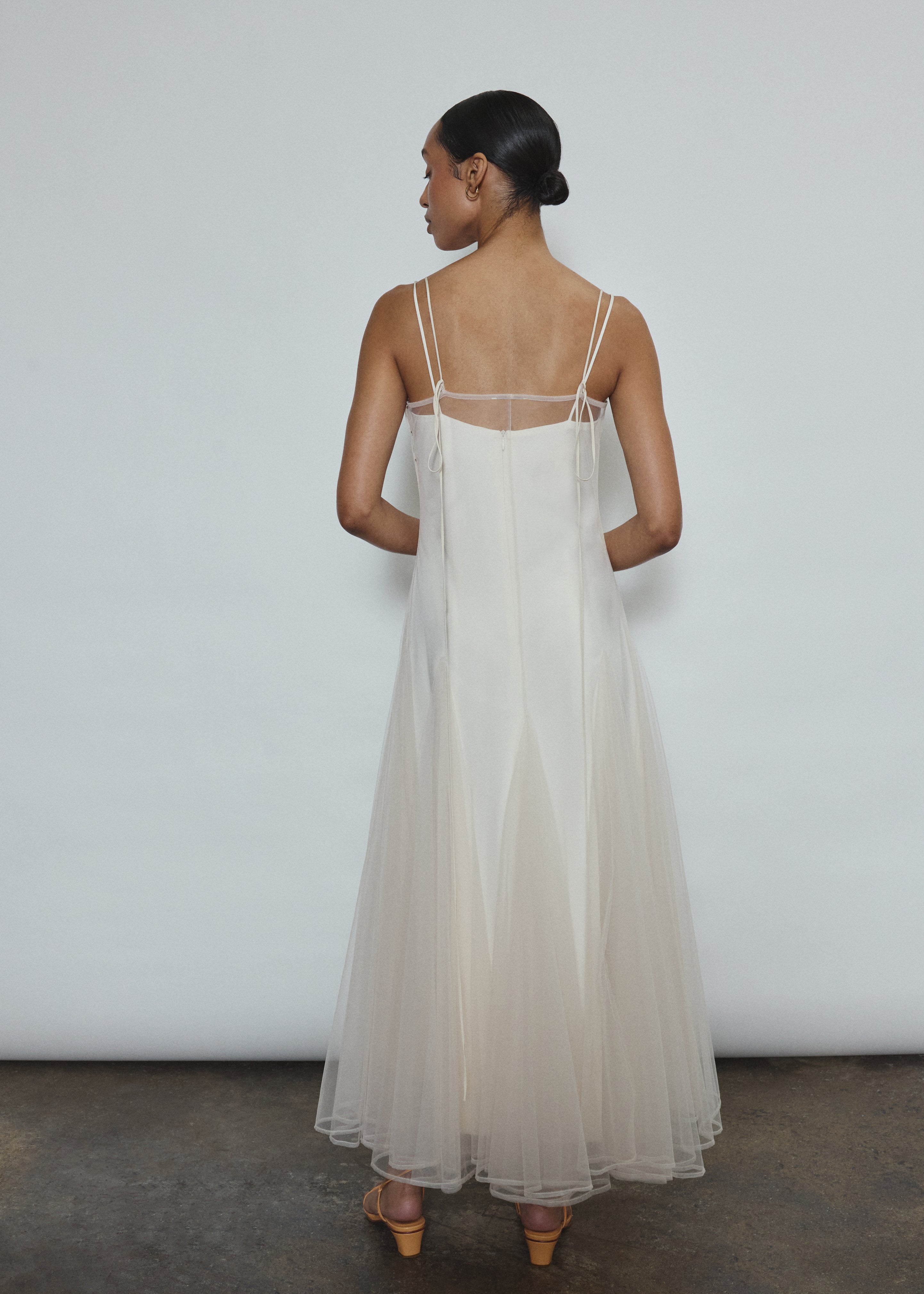 Model wearing the ivory Kamperett Camille Dress, a sheer tulle tea-length overlay featuring adjustable silk corded straps and a voluminous godet-pleated skirt.