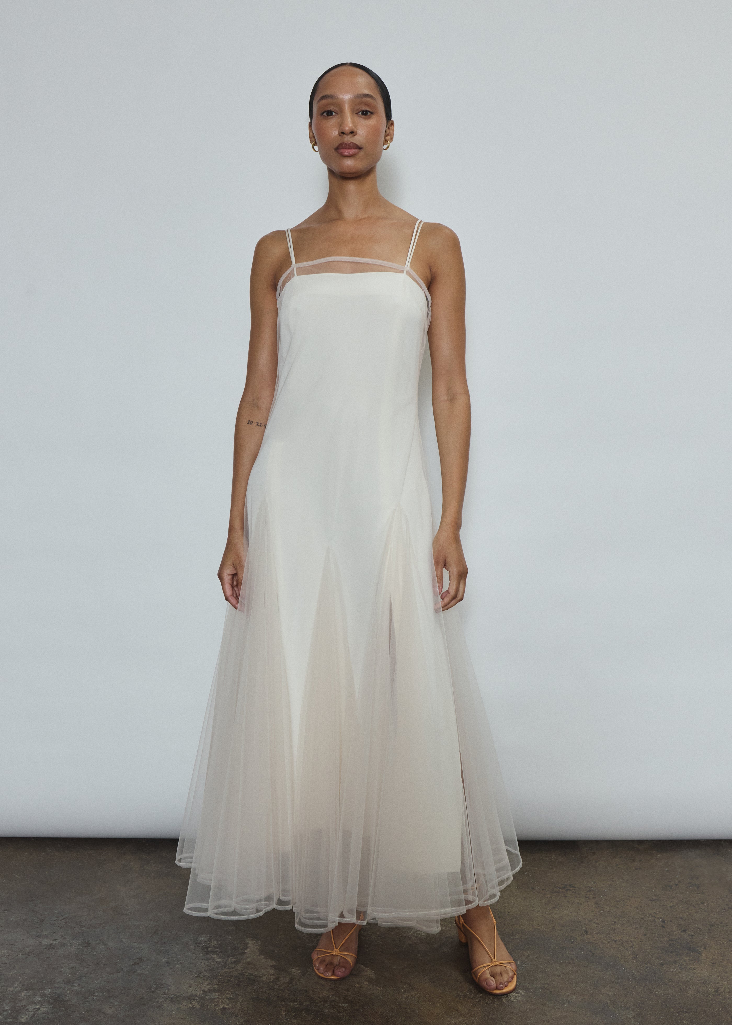 Model wearing the ivory Kamperett Camille Dress, a sheer tulle tea-length overlay featuring adjustable silk corded straps and a voluminous godet-pleated skirt.