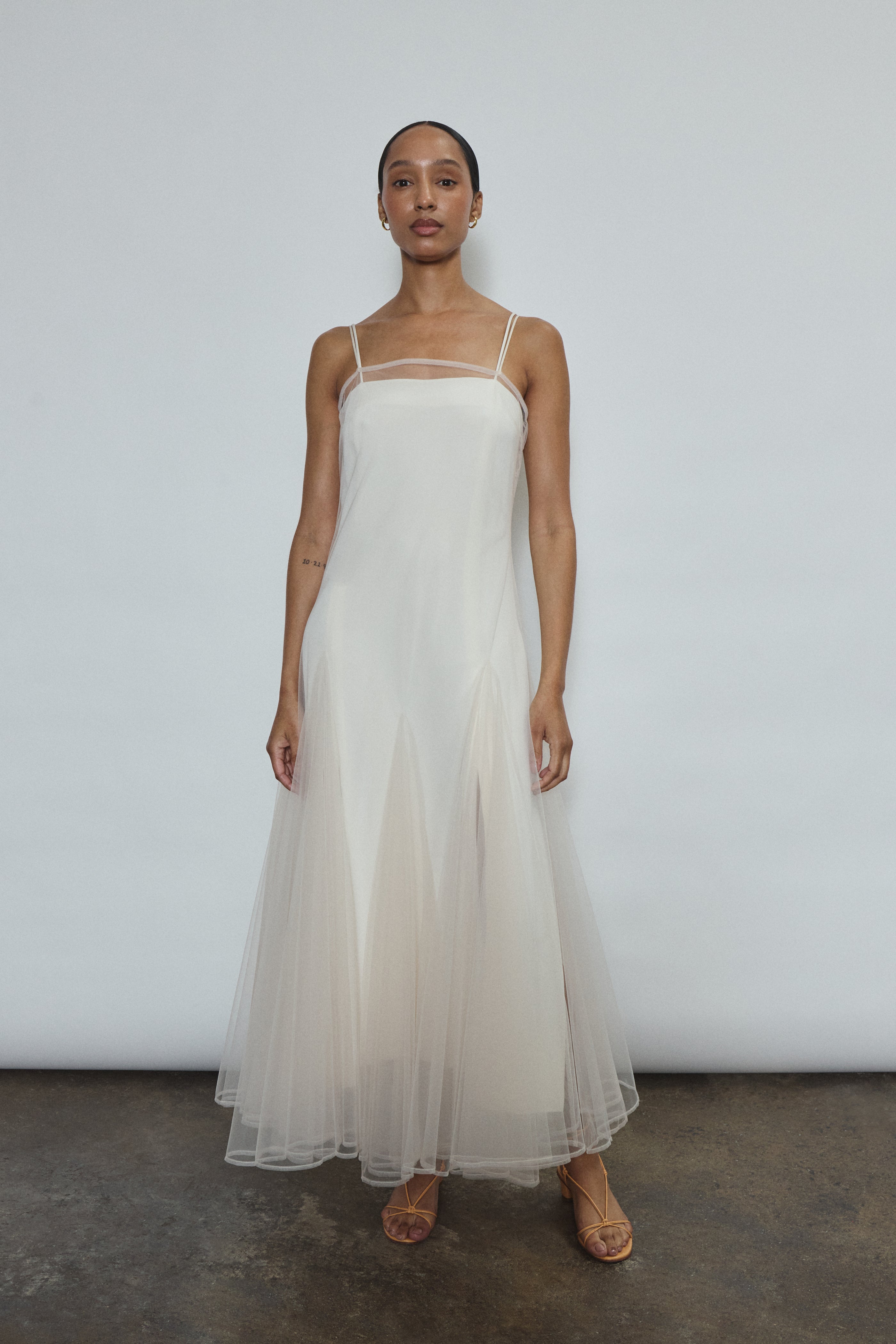 Model wearing the ivory Kamperett Camille Dress, a sheer tulle tea-length overlay featuring adjustable silk corded straps and a voluminous godet-pleated skirt.