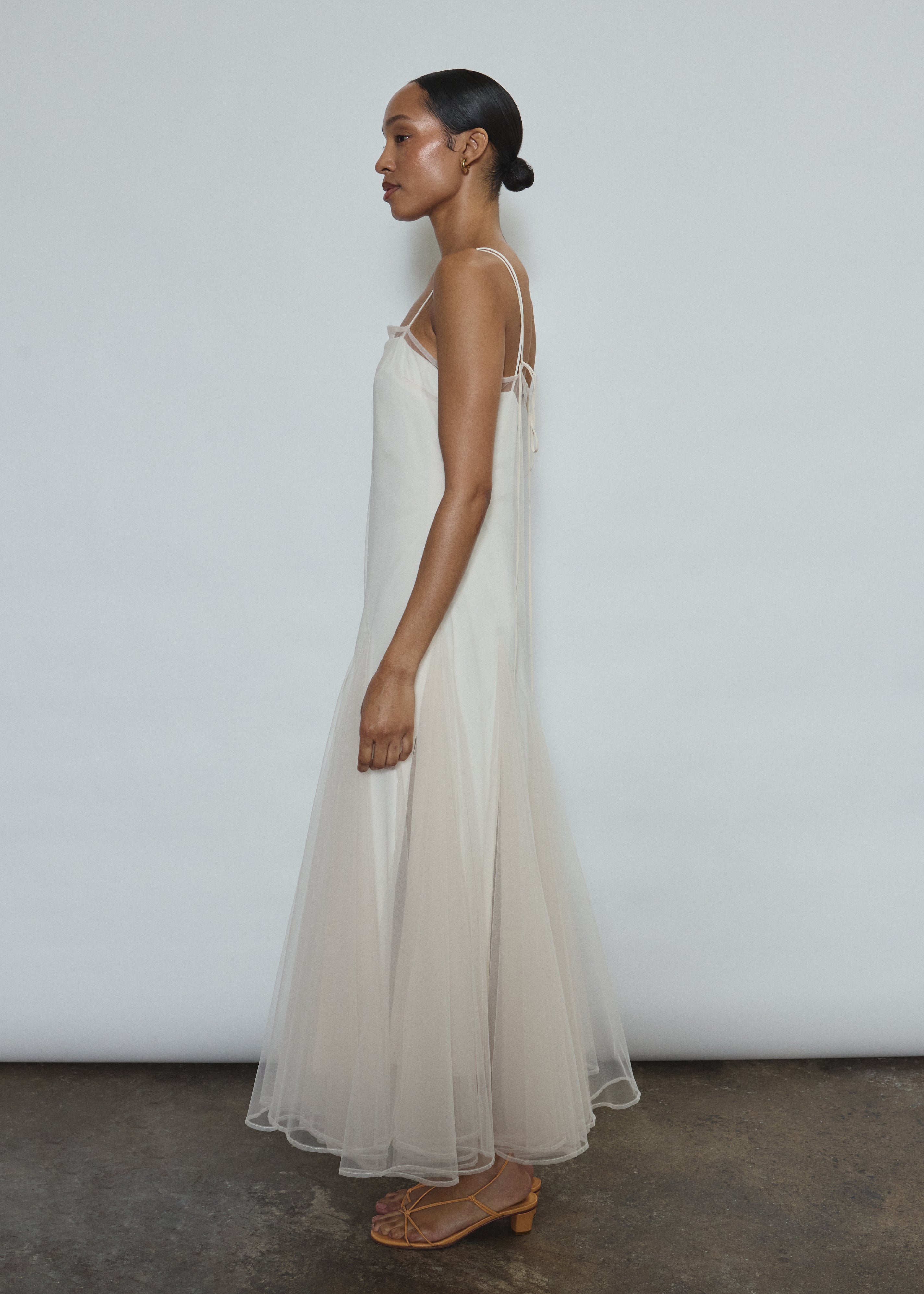 Model wearing the ivory Kamperett Camille Dress, a sheer tulle tea-length overlay featuring adjustable silk corded straps and a voluminous godet-pleated skirt.