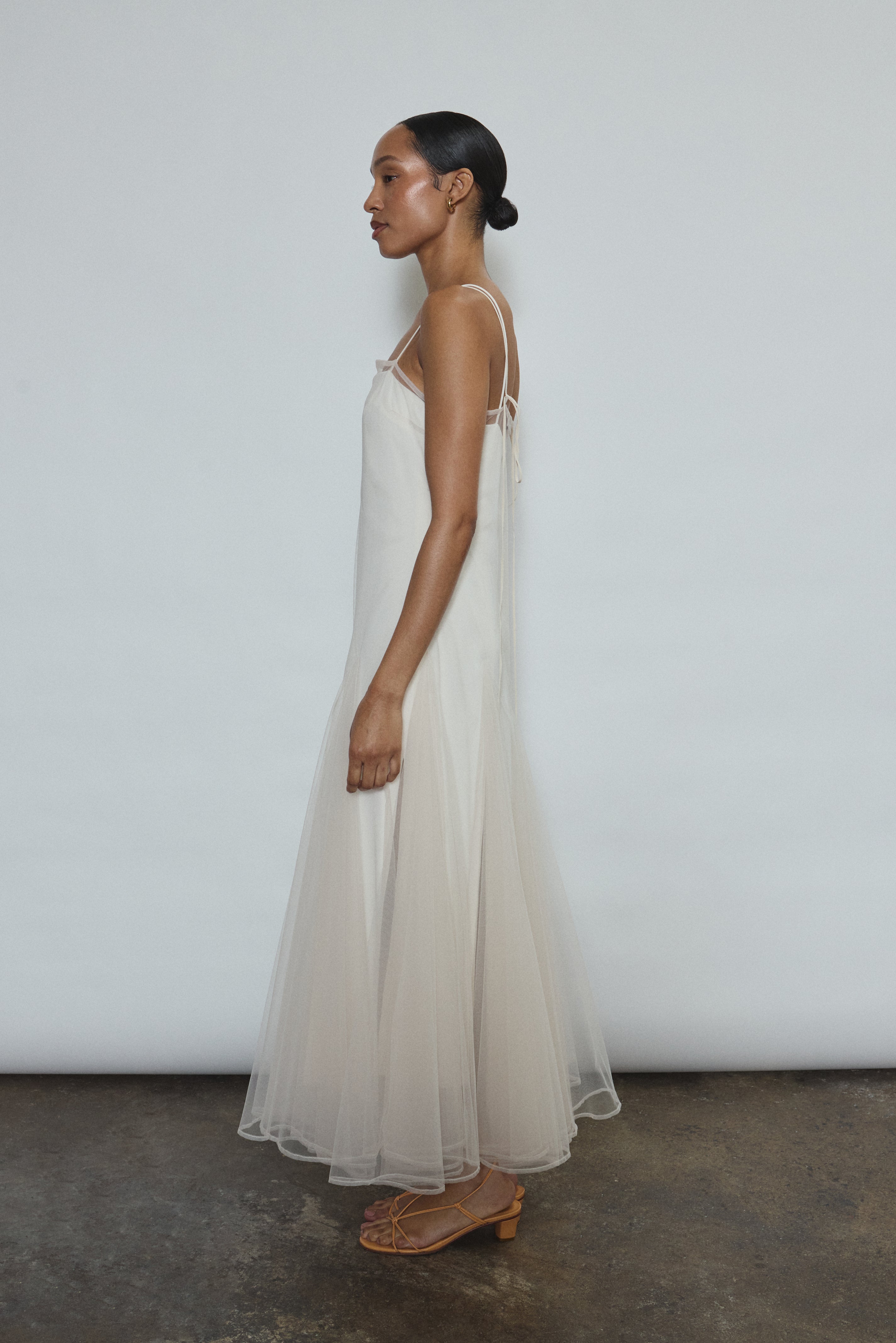 Model wearing the ivory Kamperett Camille Dress, a sheer tulle tea-length overlay featuring adjustable silk corded straps and a voluminous godet-pleated skirt.