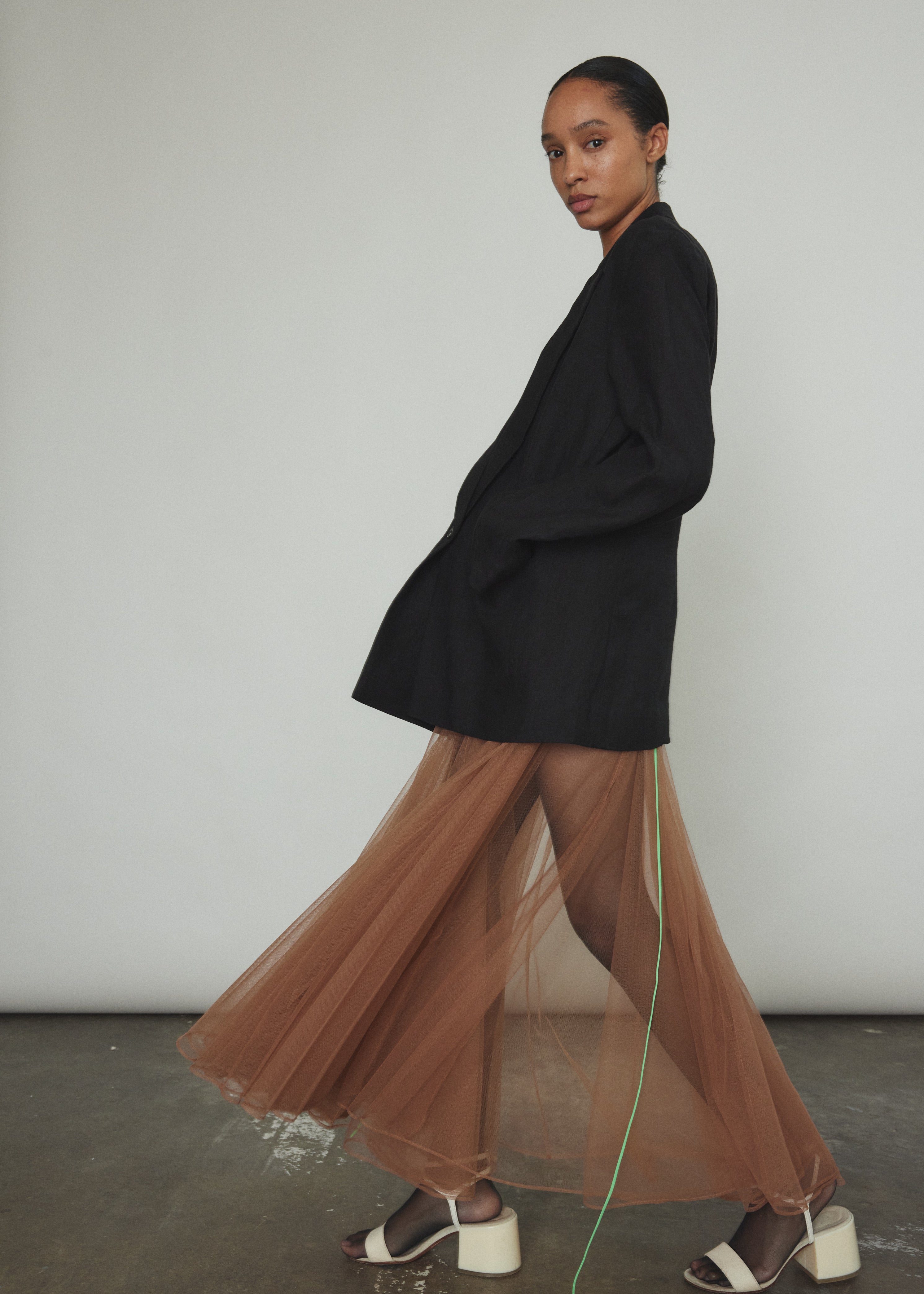 Model wearing the brown Kamperett Camille Dress, a sheer tulle tea-length overlay featuring adjustable silk corded straps and a voluminous godet-pleated skirt.