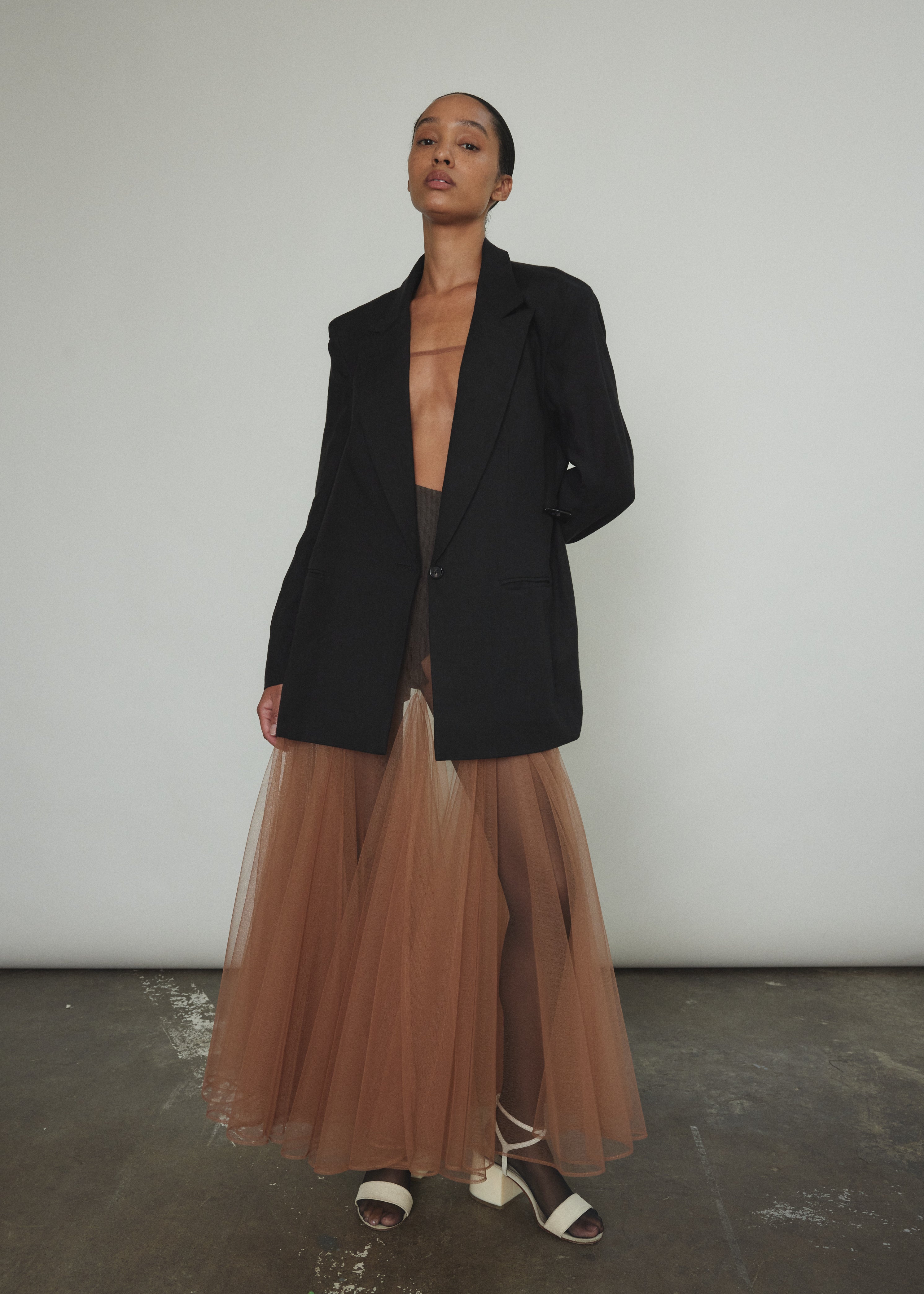 Model wearing the brown Kamperett Camille Dress, a sheer tulle tea-length overlay featuring adjustable silk corded straps and a voluminous godet-pleated skirt.