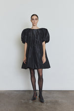 Front view of a model wearing the black silk taffeta Kamperett Eva Dress, a sculptural A-line mini with asymmetrical pleats at the neck and sleeves.