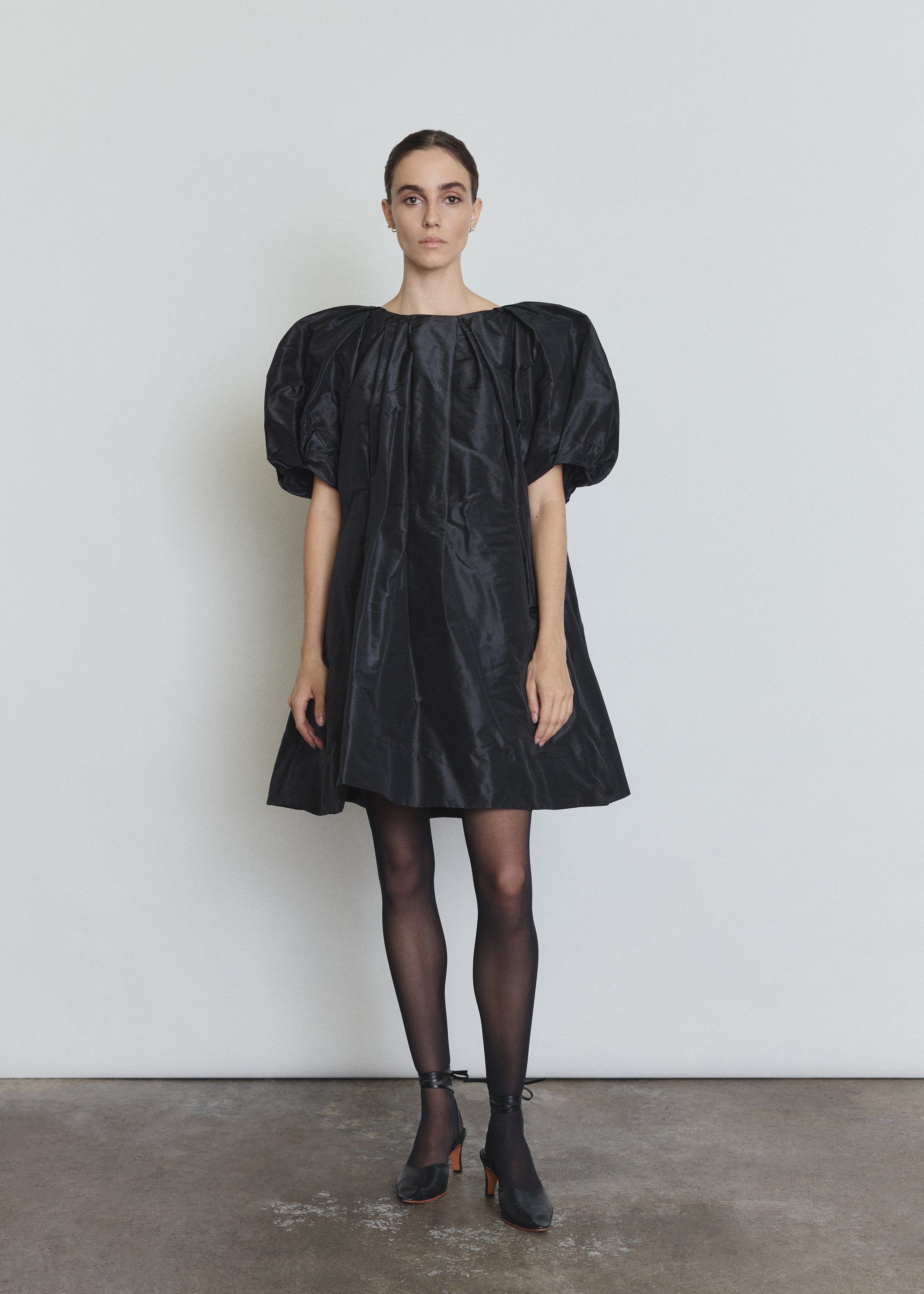 Front view of a model wearing the black silk taffeta Kamperett Eva Dress, a sculptural A-line mini with asymmetrical pleats at the neck and sleeves.
