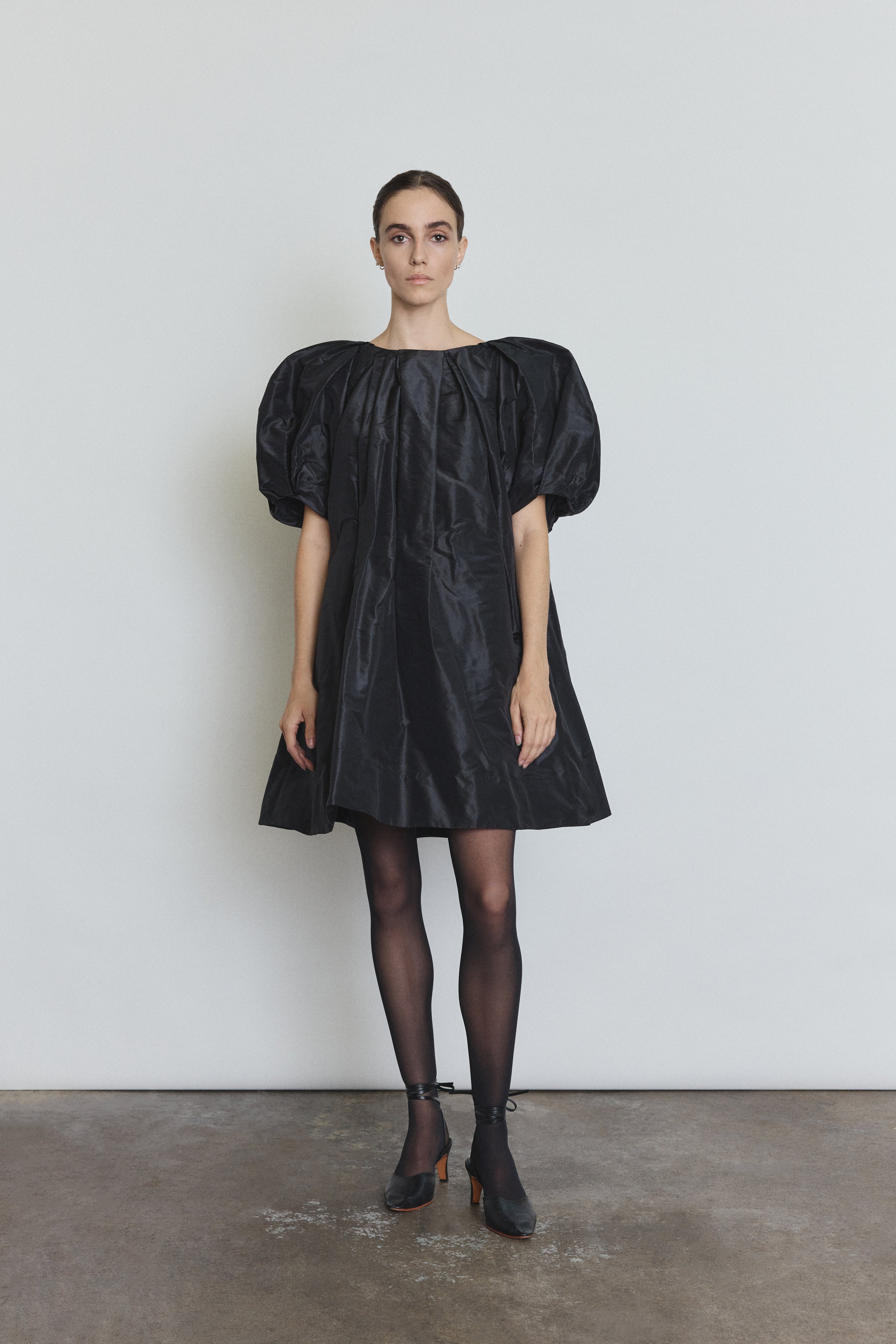 Front view of a model wearing the black silk taffeta Kamperett Eva Dress, a sculptural A-line mini with asymmetrical pleats at the neck and sleeves.