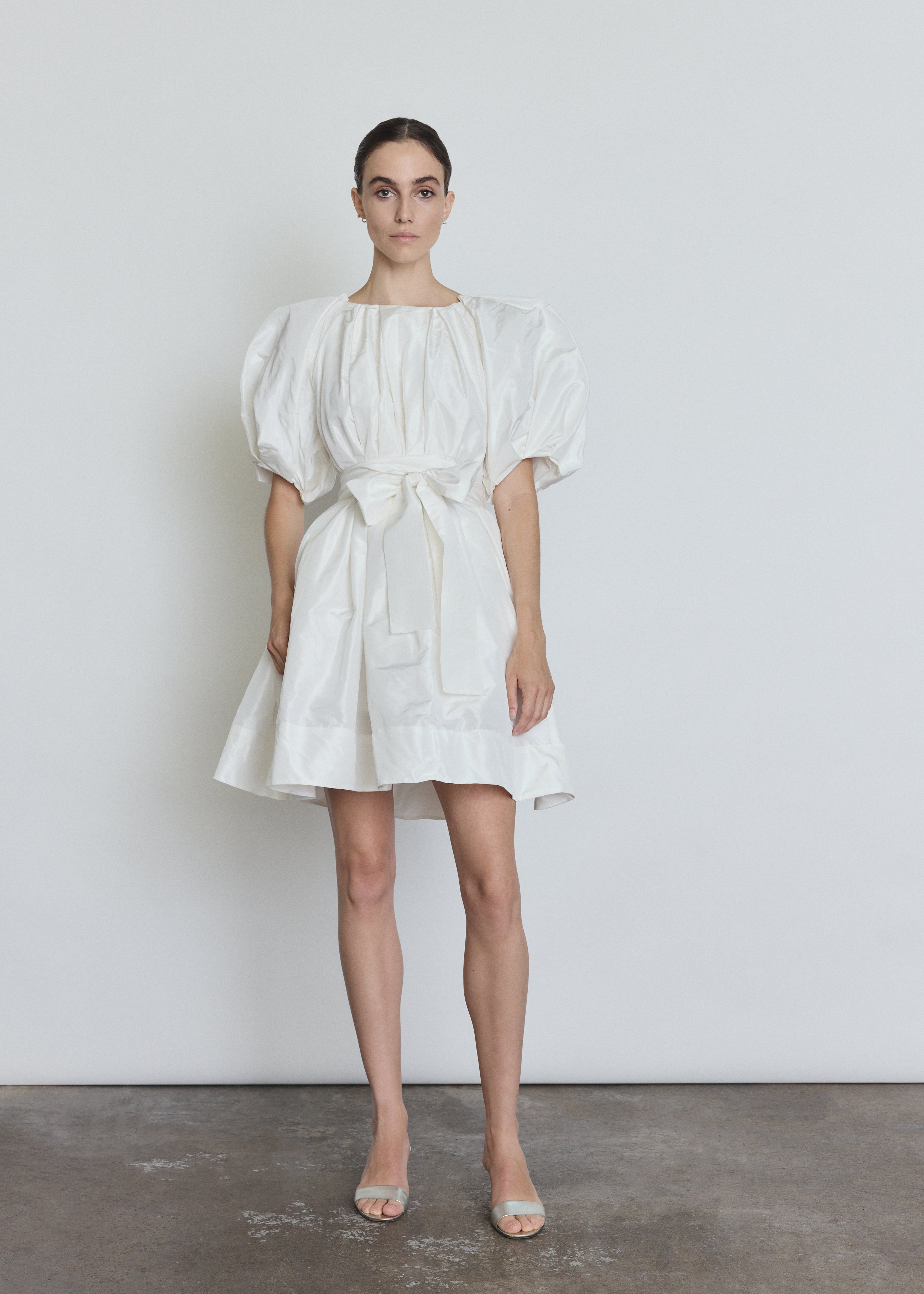 Front view of a bride wearing the white silk taffeta Kamperett Eva Dress, a sculptural bridal A-line mini with asymmetrical pleats at the neck and sleeves.