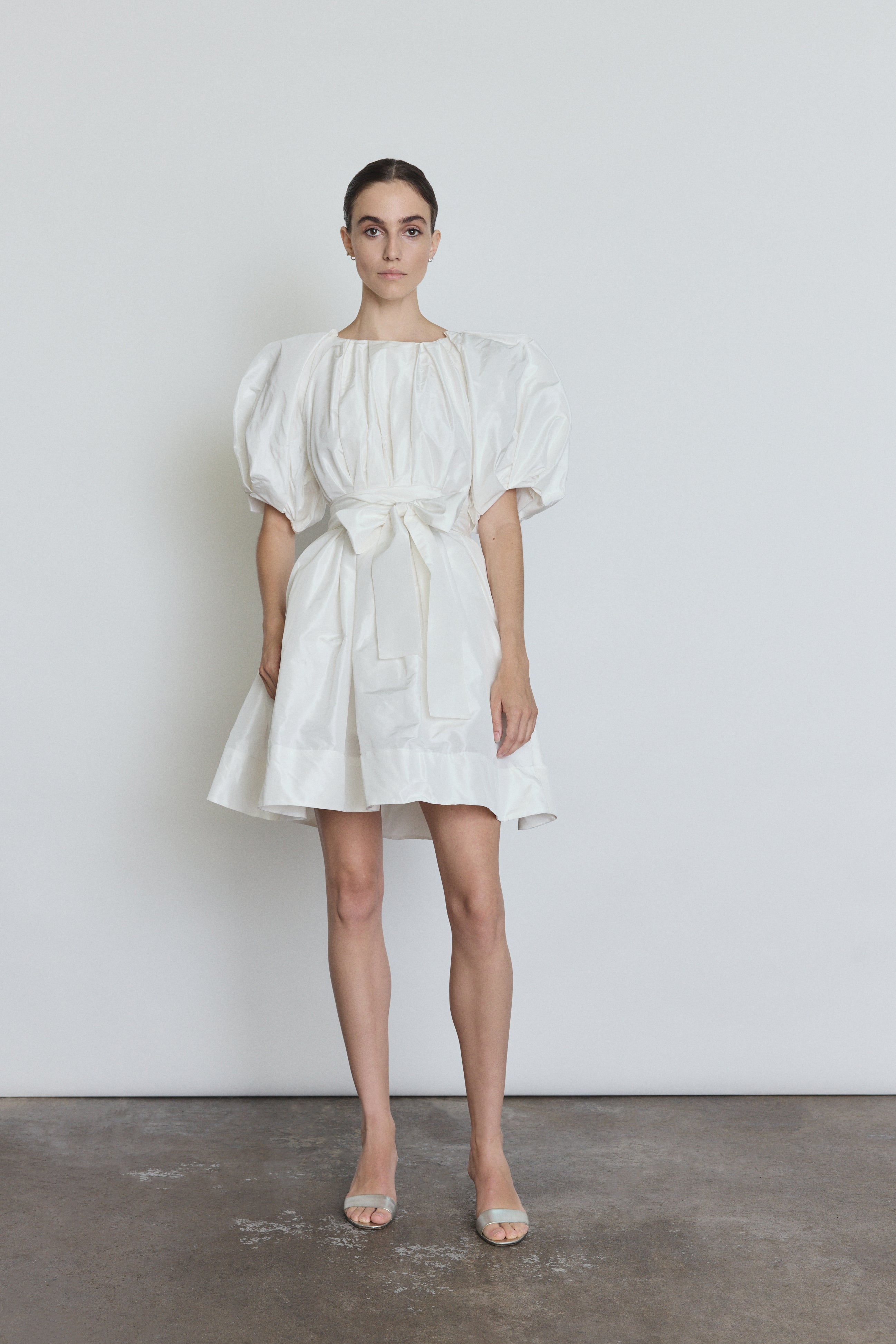 Front view of a bride wearing the white silk taffeta Kamperett Eva Dress, a sculptural bridal A-line mini with asymmetrical pleats at the neck and sleeves.