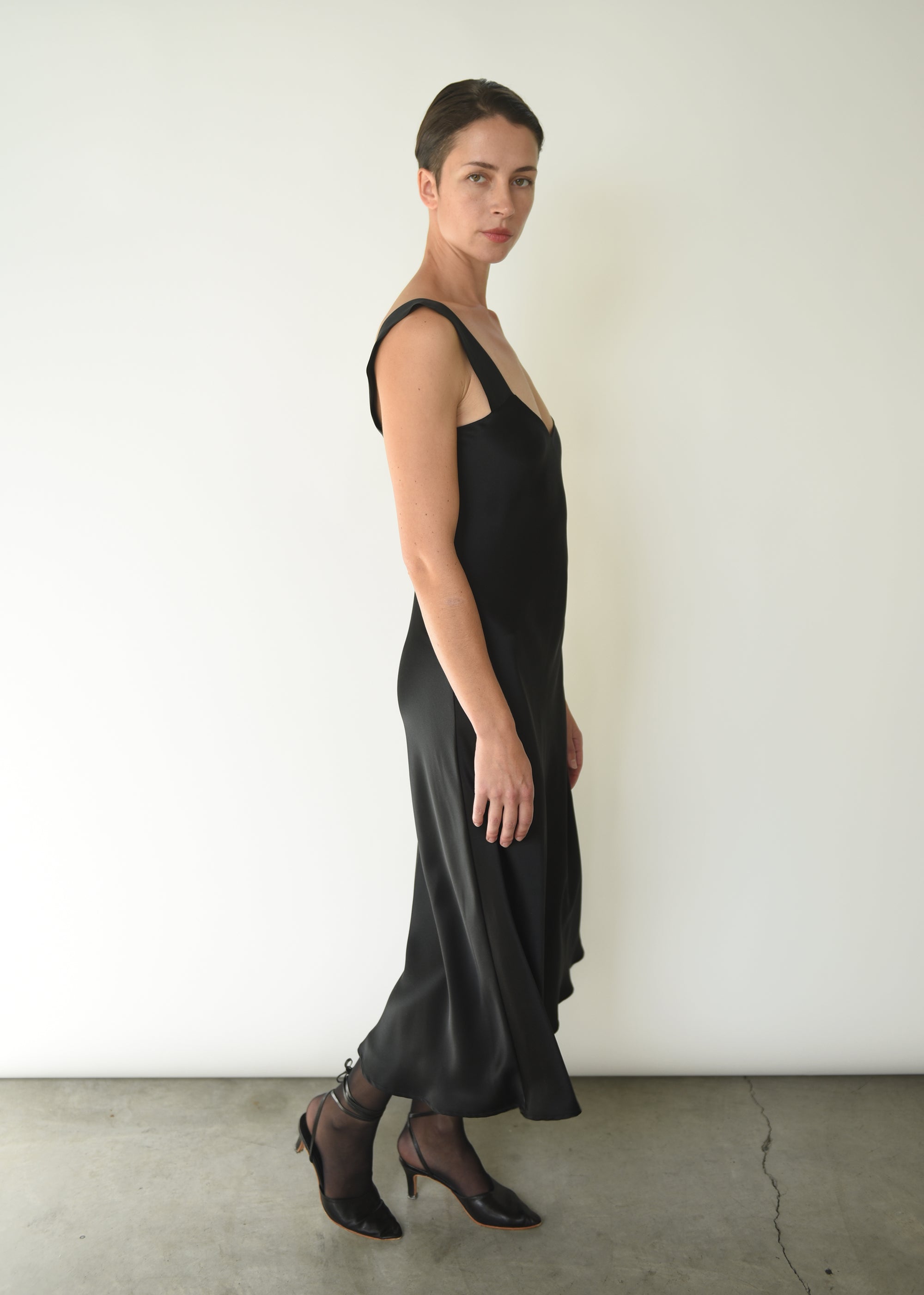 Angled view of the black silk charmeuse Kamperett Lisette Midi Slip Dress with V-neck detail.