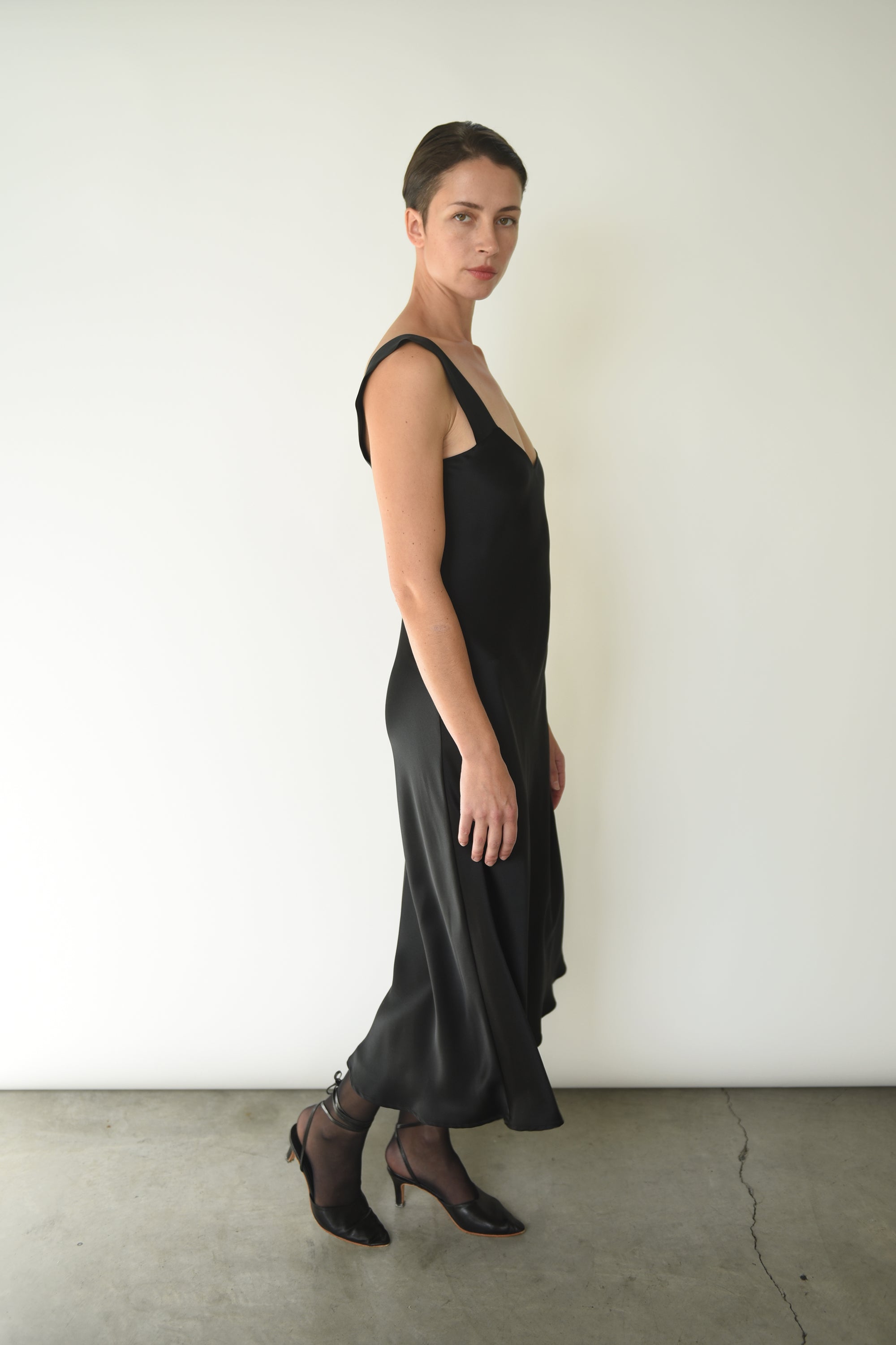 Angled view of the black silk charmeuse Kamperett Lisette Midi Slip Dress with V-neck detail.