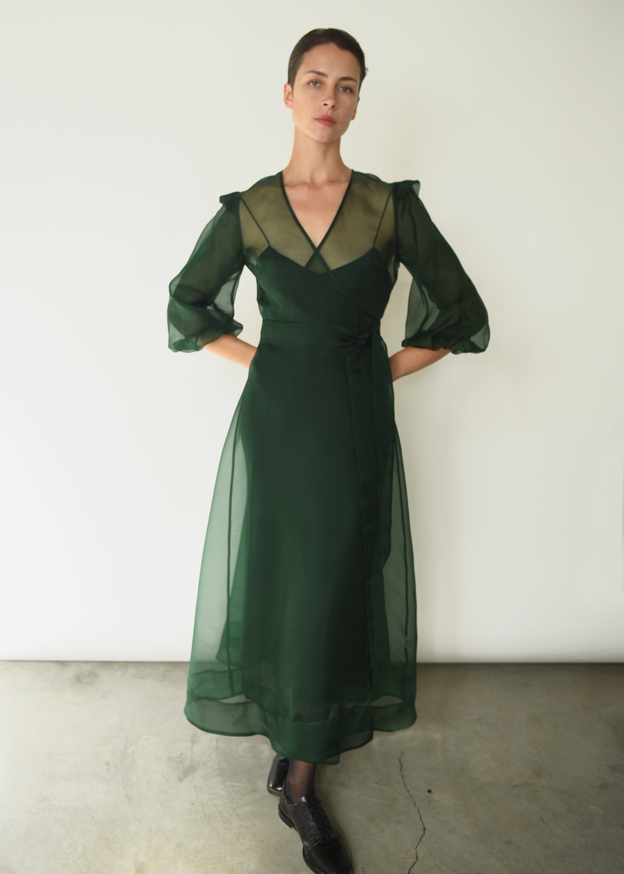 Front view of a model wearing the dark green silk organza Kamperett Loretta Midi Dress, a wrap with elbow-length sleeves and shoulder ruffle.