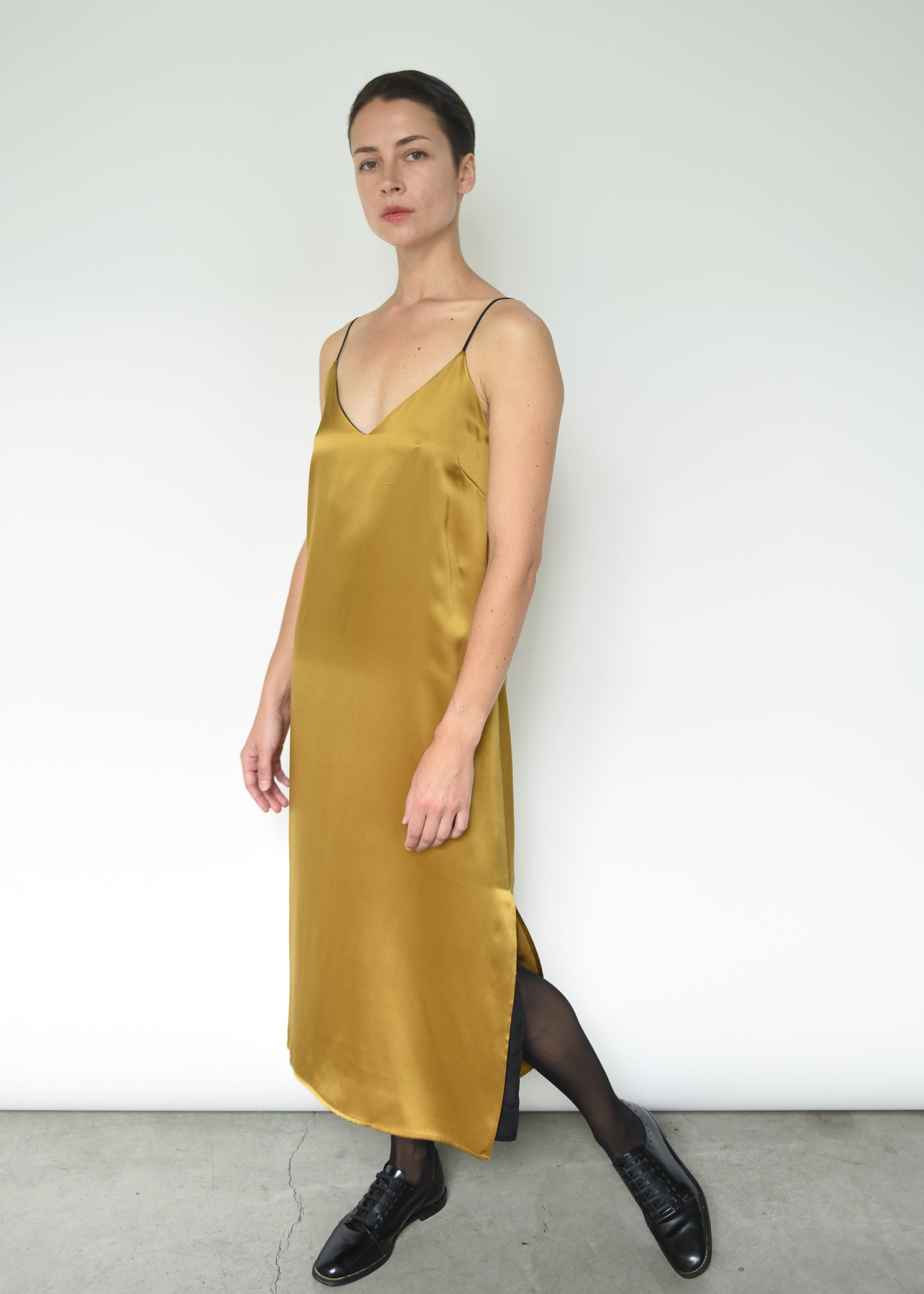 Front view of a model wearing the brass silk charmeuse Kamperett Sade Slip Dress, a straight-cut midi with corded straps.