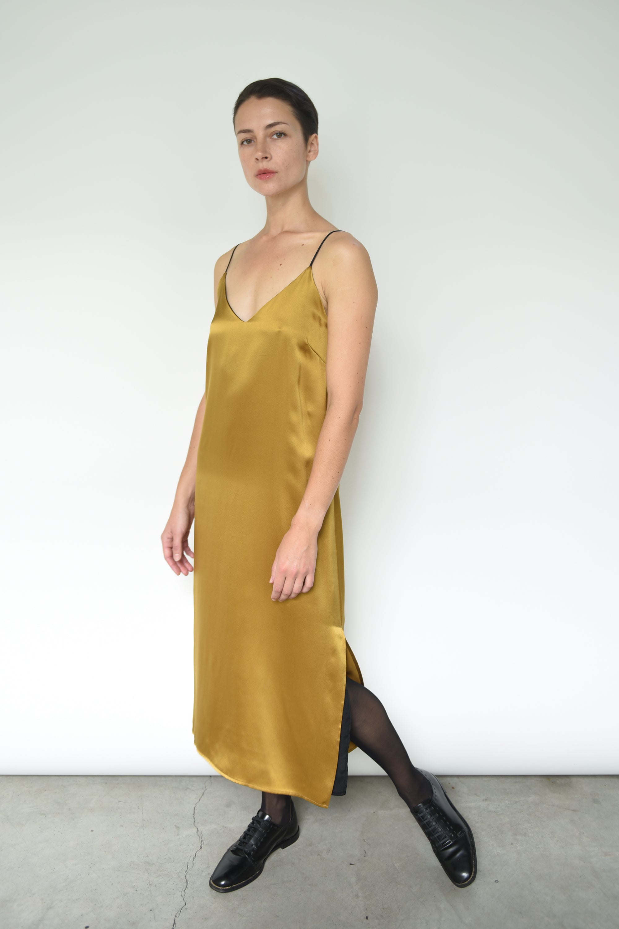 Front view of a model wearing the brass silk charmeuse Kamperett Sade Slip Dress, a straight-cut midi with corded straps.