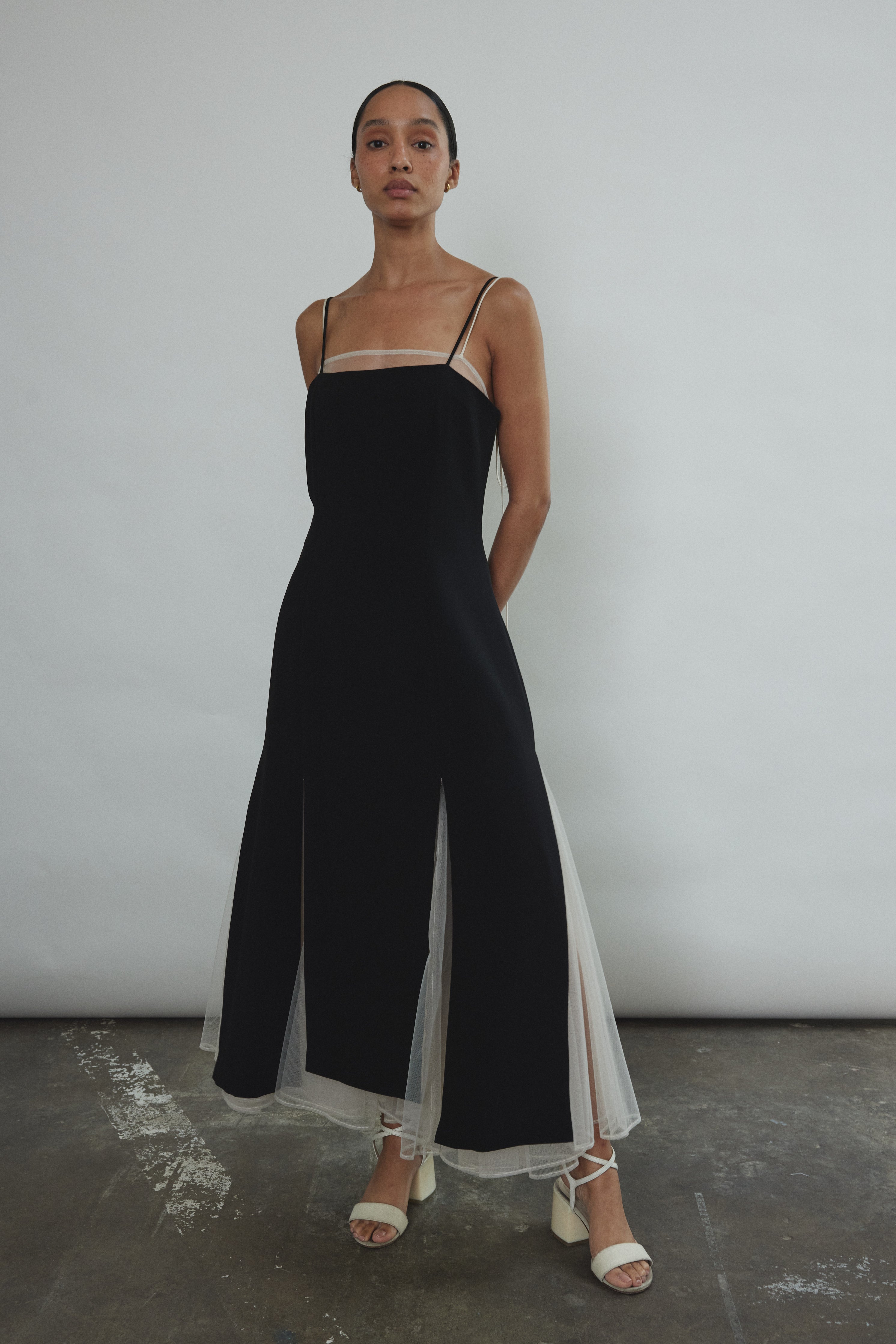 Front view of a model wearing the black crepe Kamperett Theodora Dress, a midi slip with multi-slit hemline.