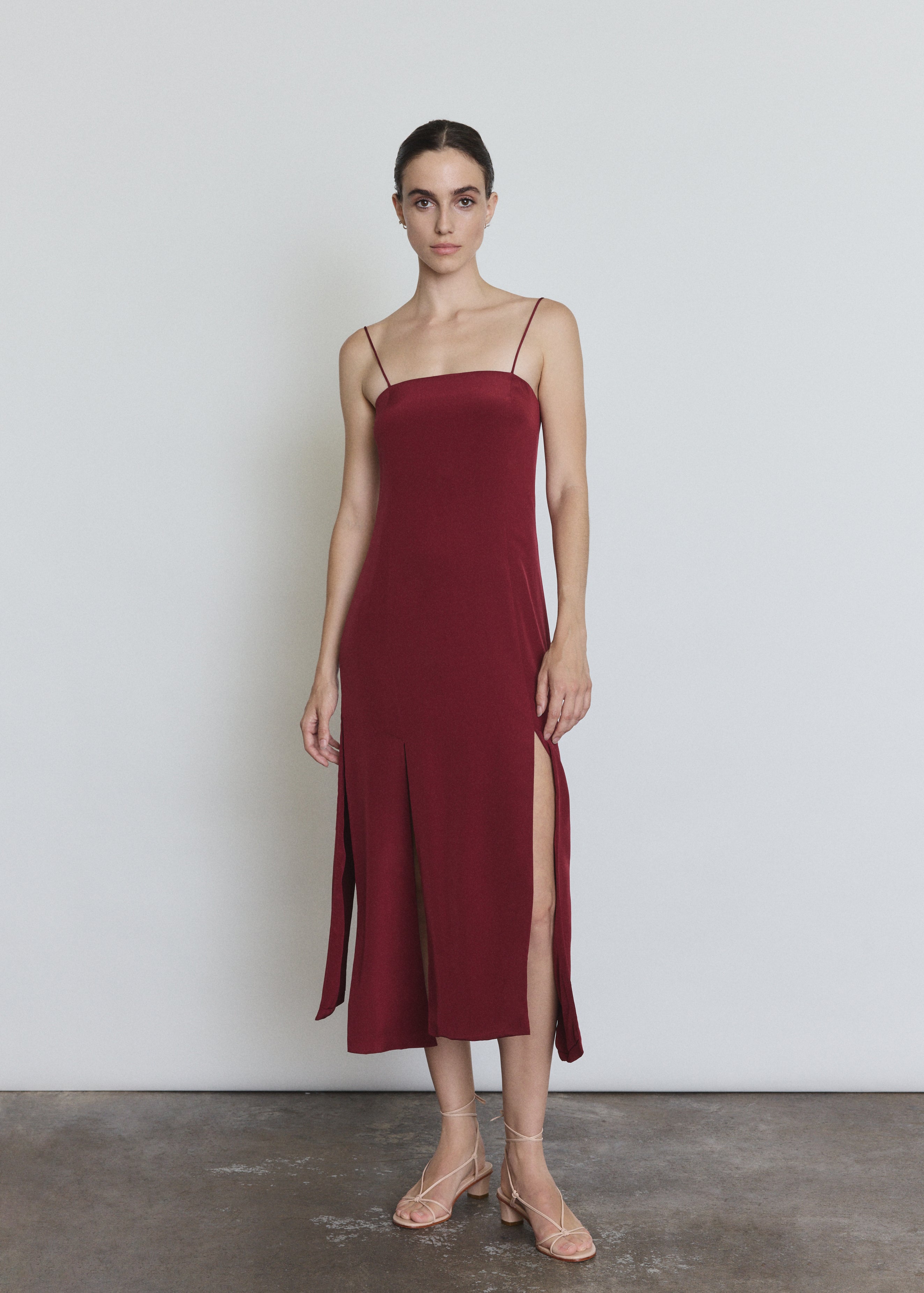 Front view of a model wearing the bordeaux silk Kamperett Theodora Dress, a sleek midi slip dress with multi-slit hemline.