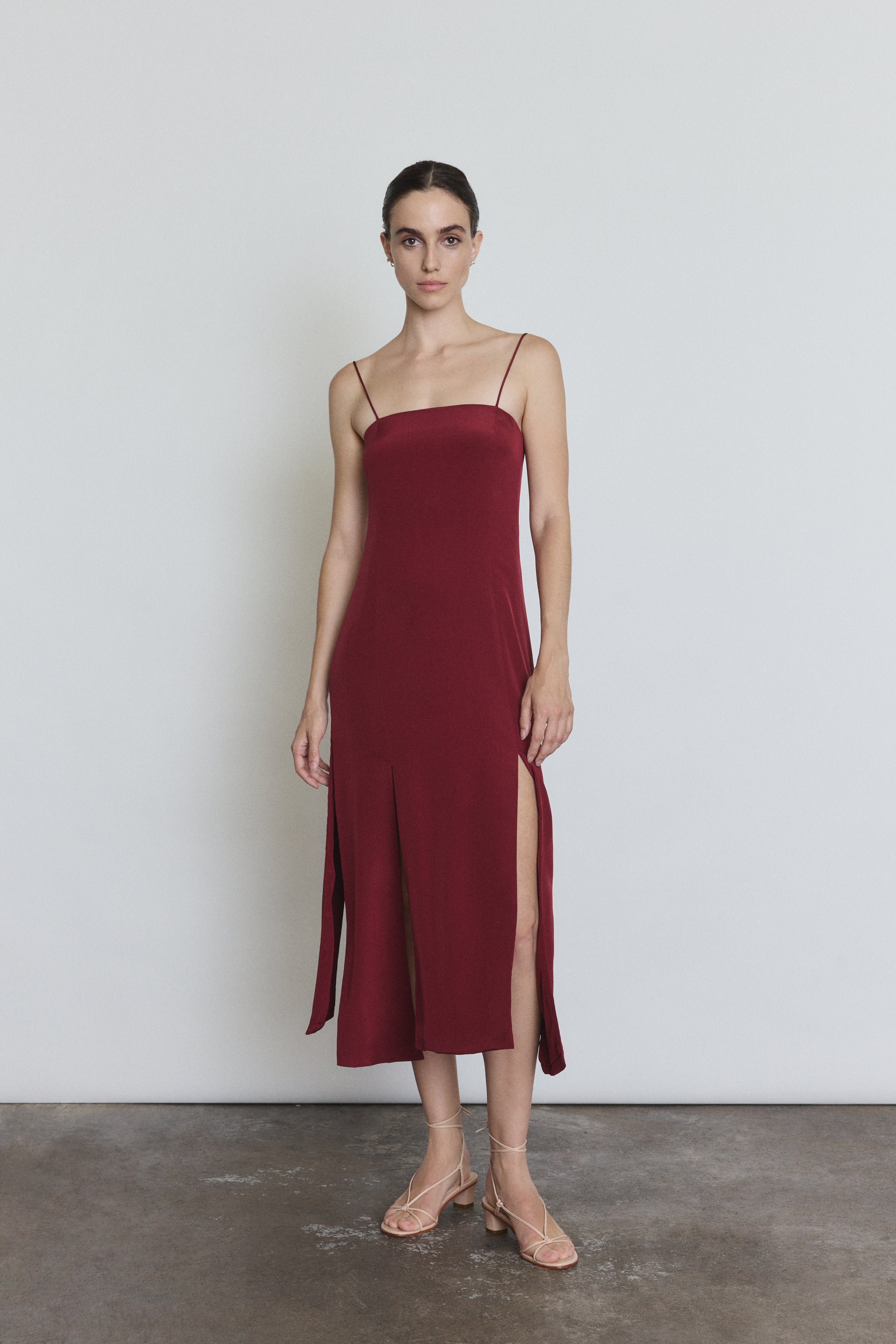 Front view of a model wearing the bordeaux silk Kamperett Theodora Dress, a sleek midi slip dress with multi-slit hemline.