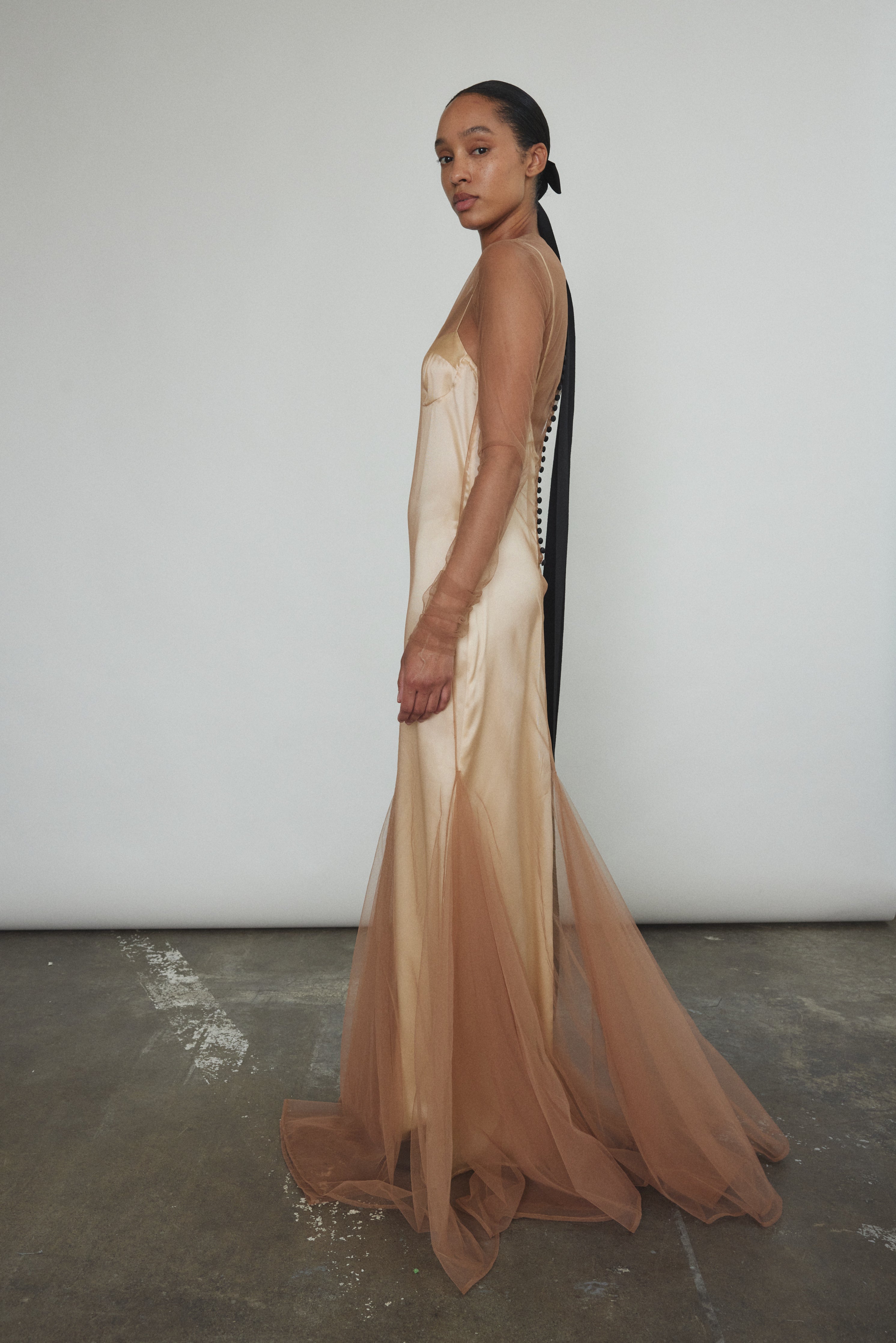 Left profile of Kamperett Violette Tulle Sheath in mocha — sleek sheath silhouette transitions into a sweeping sculptural hem, sheer tulle layer throughout.