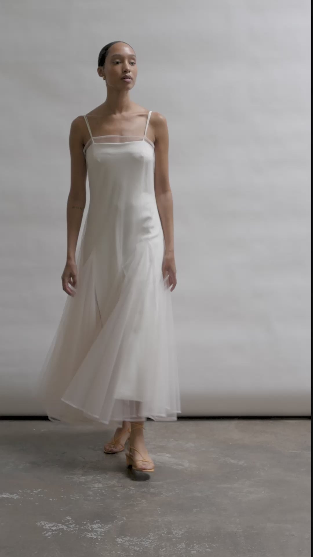 Model wearing the ivory Kamperett Camille Dress, a sheer tulle tea-length overlay featuring adjustable silk corded straps and a voluminous godet-pleated skirt.