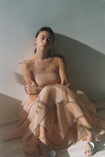 Front view of a model wearing the blush silk organza Kamperett Laurence Midi Dress, a tiered midi with corded shoulder ties and black pearl edge trim.